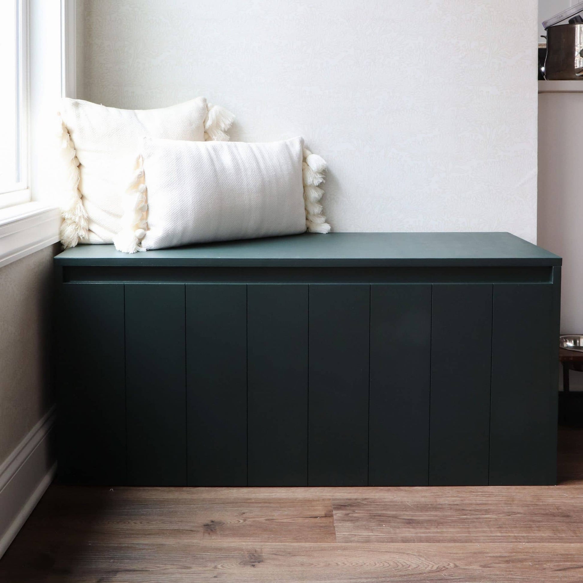 Green storage bench with white cushions in a room with wooden flooring and a window.